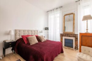 A cozy bedroom featuring a tufted bed with red bedding, elegant pillows, and a classic fireplace with a decorative mirror above. Natural light pours in through large windows with sheer curtains.