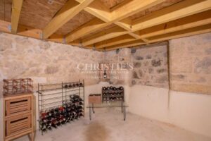 A wine cellar with stone walls and a wooden ceiling, featuring wine racks filled with bottles and a few small wooden cabinets.