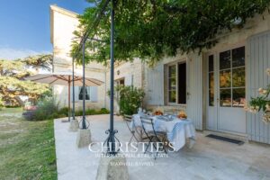Une terrasse en pierre avec une table à manger, ornée d'une nappe blanche, se trouve devant une maison avec des volets bleus et des arbres ombrageux.