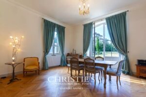Une salle à manger élégante avec de grandes fenêtres, des rideaux bleus, et un parquet en bois. Une table en bois entourée de chaises se trouve au centre, éclairée par un lustre au plafond.