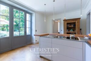 Une cuisine moderne avec des armoires blanches et un îlot central, dotée de grandes fenêtres offrant une vue sur un jardin.