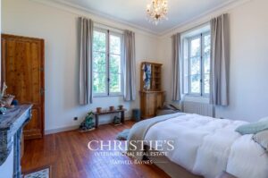 Une chambre lumineuse avec un sol en bois, un lit double confortable, deux grandes fenêtres avec des rideaux beiges et une armoire en bois.