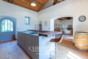 Une cuisine moderne et lumineuse avec un îlot central, un tabouret, et un tonneau en bois, reliée à un salon ouvert.