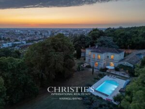 Vue aérienne d'une maison avec piscine au crépuscule, entourée de verdure et donnant sur une ville à l'horizon.