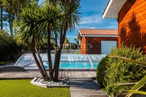 Jardin avec piscine couverte, palmiers et maison en bois sous un ciel bleu.