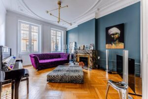 Un salon élégant avec un canapé violet, une cheminée ornée, des murs bleu-gris et une peinture au-dessus. Un piano et des meubles modernes complètent l'espace.