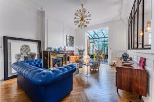 Salon élégant avec un canapé bleu, parquet en bois, cheminée décorative et grande baie vitrée laissant entrer la lumière.