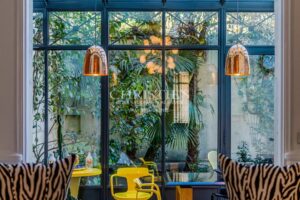 Une pièce lumineuse avec de grandes fenêtres montrant un jardin verdoyant, des lampes suspendues en métal et des chaises jaunes.