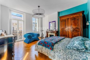 Une chambre lumineuse avec un grand lit, une armoire en bois, un fauteuil bleu et des fenêtres donnant sur l'extérieur.