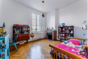 Une chambre d'enfant lumineuse avec un parquet en bois, un lit rose, des étagères remplies de livres et de jouets, et un bureau avec des chaises.