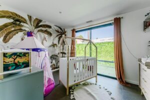 A brightly lit nursery featuring a white crib, a colorful play tent, and nature-themed wall decor with palm leaves.