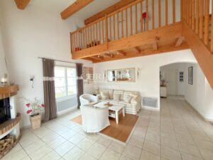 Un salon lumineux avec un plafond en bois, un canapé à carreaux, une table basse, et une cheminée d'angle à côté d'une grande fenêtre.